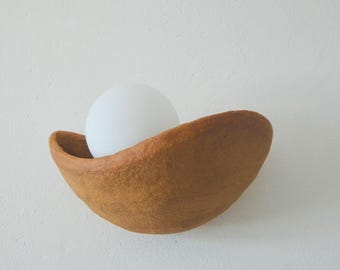 May include: A wall-mounted light fixture featuring a textured, bowl-shaped base in a warm, earthy brown tone. Nestled within the curved, organic form of the base is a smooth, spherical white glass globe that serves as the light source.