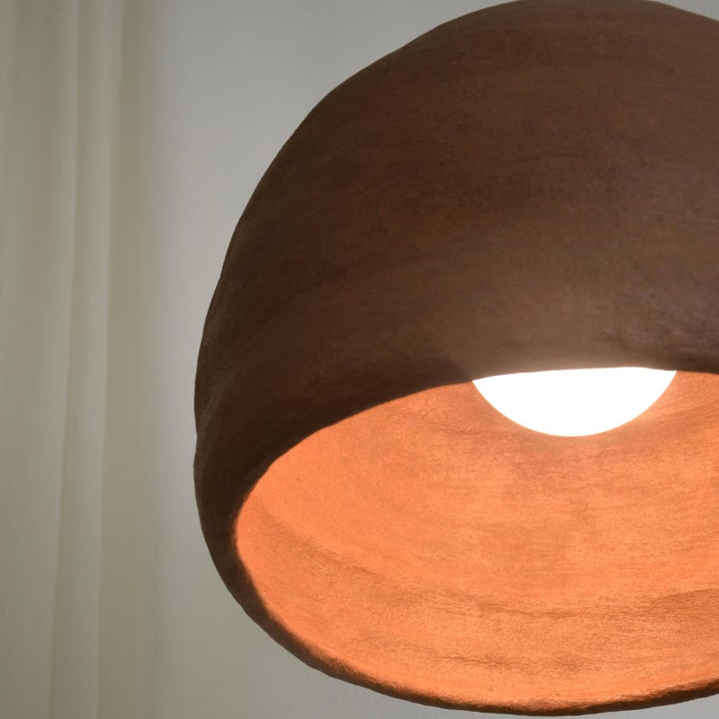 May include: A brown, textured pendant light with a warm, orange interior. The light fixture has a rounded, organic shape and a bright, white light bulb is visible inside. The background is a soft, off-white color.