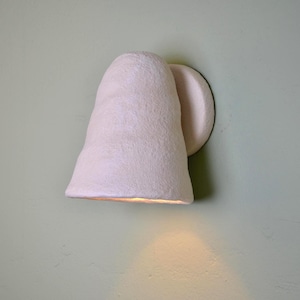 May include: A wall-mounted light fixture with a textured, off-white, conical shade. The lamp features a rounded base plate attached to the wall, with the shade angled downward to cast a warm, soft glow onto the surface directly below the fixture.