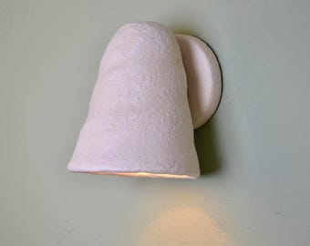 Puede incluir: Un aplique de pared con una pantalla cónica texturizada de color blanco roto. La lámpara cuenta con una placa base redondeada fijada a la pared, con la pantalla inclinada hacia abajo para proyectar una luz cálida y suave sobre la superficie inferior.