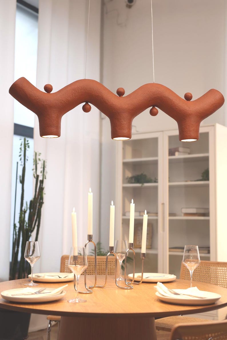 May include: A terracotta-colored pendant light fixture with a wavy, branch-like structure featuring three downward-facing lights and small spherical accents, suspended above a round wooden dining table with a metal candelabra and place settings.