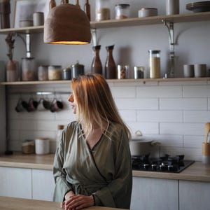 May include: A kitchen scene with a woman wearing a green robe standing near a wooden countertop. A brown, bell-shaped pendant light hangs above. Shelves hold jars, bottles, and mugs. The kitchen has white tile and light-colored cabinets.