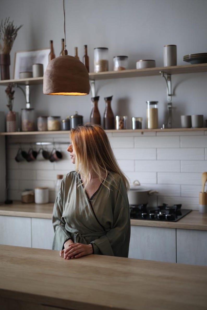 May include: A kitchen scene with a woman wearing a green robe standing near a wooden countertop. A brown, bell-shaped pendant light hangs above. Shelves hold jars, bottles, and mugs. The kitchen has white tile and light-colored cabinets.