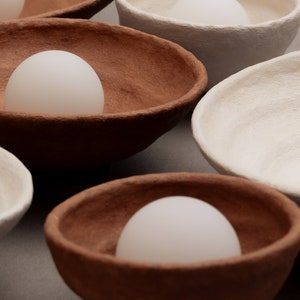 May include: A collection of textured, hand-formed bowls in earthy terracotta and off-white tones. Each bowl features an irregular, organic rim and contains a smooth, opaque white spherical object resting in the centre of its basin.