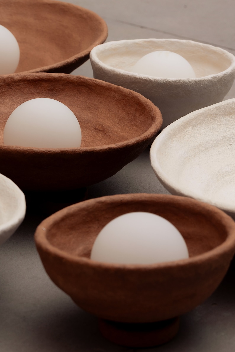 May include: A collection of textured, hand-formed bowls in earthy terracotta and off-white tones. Each bowl features an irregular, organic rim and contains a smooth, opaque white spherical object resting in the centre of its basin.