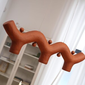 May include: A terracotta-colored hanging light fixture with a wavy, organic, branch-like structure. The surface has a textured, matte finish and features several small, spherical protrusions along its length, suspended by thin wires.