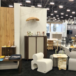 May include: A white, fluffy, modern chair with a unique shape sits in a room with a wooden cabinet, a striped chair, and a table with a patterned top. The room has a white wall with a brown wooden panel behind the cabinet.