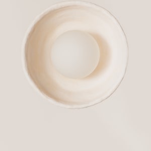 May include: A top-down view of a circular, off-white bowl with a textured, matte surface. Centered inside the bowl is a smooth, opaque, spherical object in a lighter shade of white, creating a clean, monochromatic composition with soft shadows.