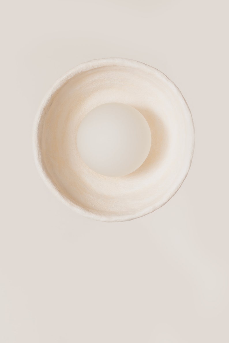 May include: A top-down view of a circular, off-white bowl with a textured, matte surface. Centered inside the bowl is a smooth, opaque, spherical object in a lighter shade of white, creating a clean, monochromatic composition with soft shadows.