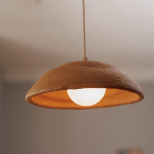 Pode incluir: Uma lumin&aacute;ria pendente com uma abajur castanho texturizado e uma l&acirc;mpada redonda e brilhante. O abajur tem um interior laranja quente. A luz pende de um fino cabo bege.