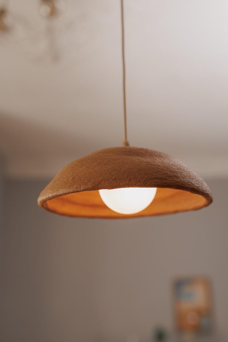 Pode incluir: Uma lumin&aacute;ria pendente com uma abajur castanho texturizado e uma l&acirc;mpada redonda e brilhante. O abajur tem um interior laranja quente. A luz pende de um fino cabo bege.