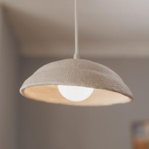 Pode incluir: Uma lumin&aacute;ria pendente com uma sombra texturizada em branco sujo e uma l&acirc;mpada redonda e brilhante. A luz pende de um cabo branco, criando um brilho suave e ambiente. O design da lumin&aacute;ria &eacute; simples e moderno.