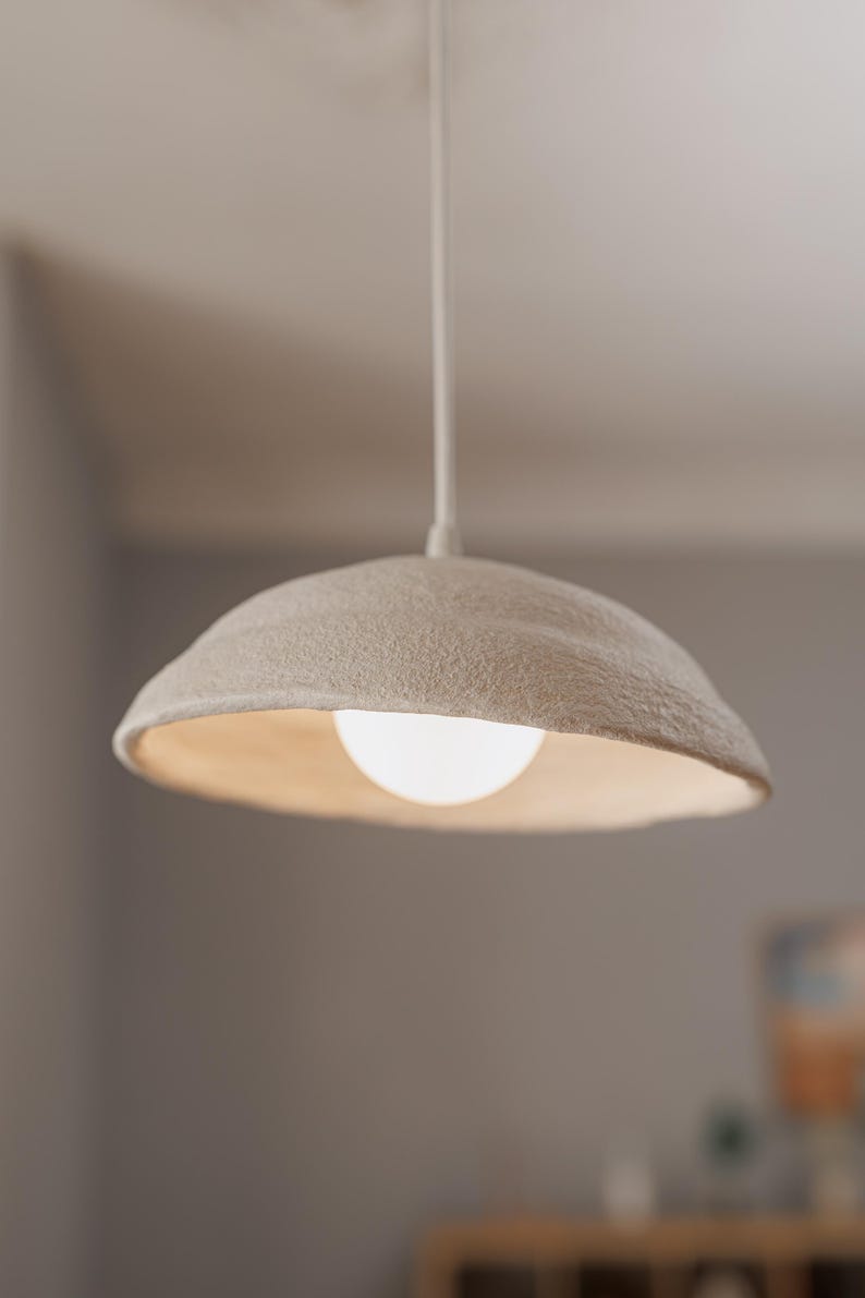 Pode incluir: Uma lumin&aacute;ria pendente com uma sombra texturizada em branco sujo e uma l&acirc;mpada redonda e brilhante. A luz pende de um cabo branco, criando um brilho suave e ambiente. O design da lumin&aacute;ria &eacute; simples e moderno.