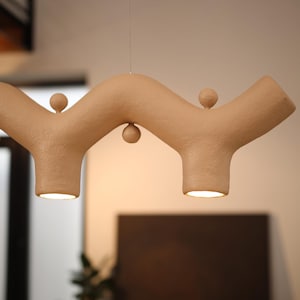 May include: A beige pendant light with a textured, wavy, branch-like structure featuring two downward-facing light openings and three small spherical accents positioned along the top and bottom curves of the horizontal fixture.