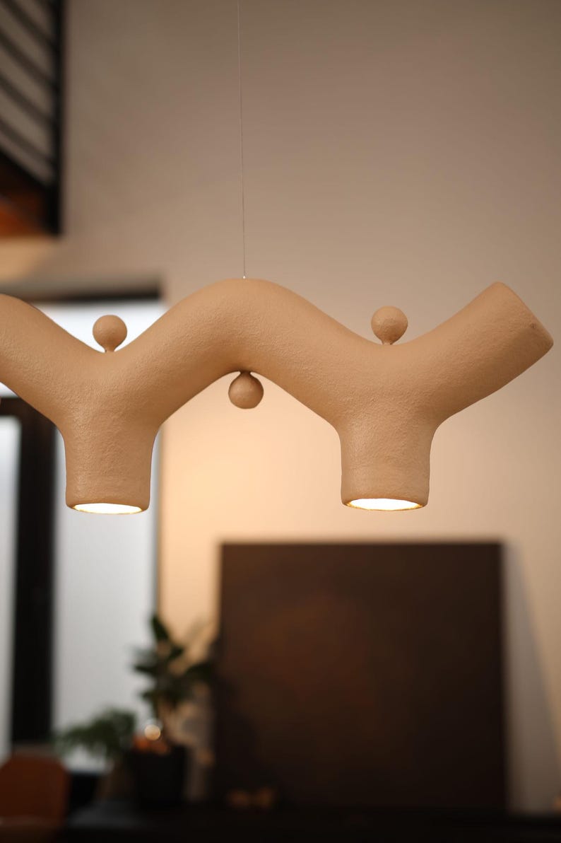 May include: A beige pendant light with a textured, wavy, branch-like structure featuring two downward-facing light openings and three small spherical accents positioned along the top and bottom curves of the horizontal fixture.