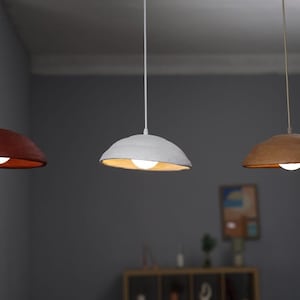 Pode incluir: Tr&ecirc;s lumin&aacute;rias pendentes com um design org&acirc;nico &uacute;nico. Uma &eacute; vermelho escuro, outra branca e a terceira marrom quente. Cada uma tem uma &uacute;nica l&acirc;mpada, criando um brilho suave e convidativo. As luzes pendem do teto.