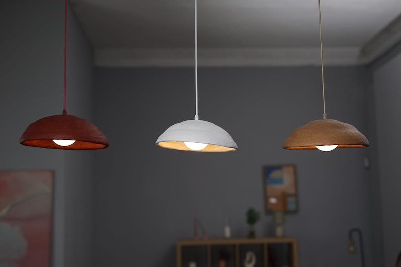 Pode incluir: Tr&ecirc;s lumin&aacute;rias pendentes com um design org&acirc;nico &uacute;nico. Uma &eacute; vermelho escuro, outra branca e a terceira marrom quente. Cada uma tem uma &uacute;nica l&acirc;mpada, criando um brilho suave e convidativo. As luzes pendem do teto.