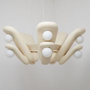 Op de afbeelding: Een beige, organisch gevormde kroonluchter met zes witte bolvormige lampen. De kroonluchter hangt aan dunne witte koorden aan het plafond.