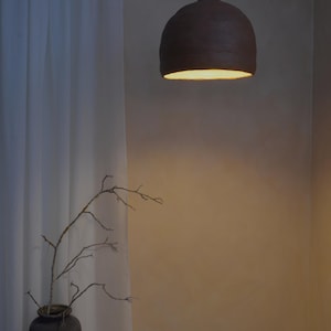 May include: A dark brown, dome-shaped pendant light fixture hangs from the ceiling. The light fixture is illuminated from within, casting a warm glow. A vase with branches is in the foreground, and a white curtain is to the left.