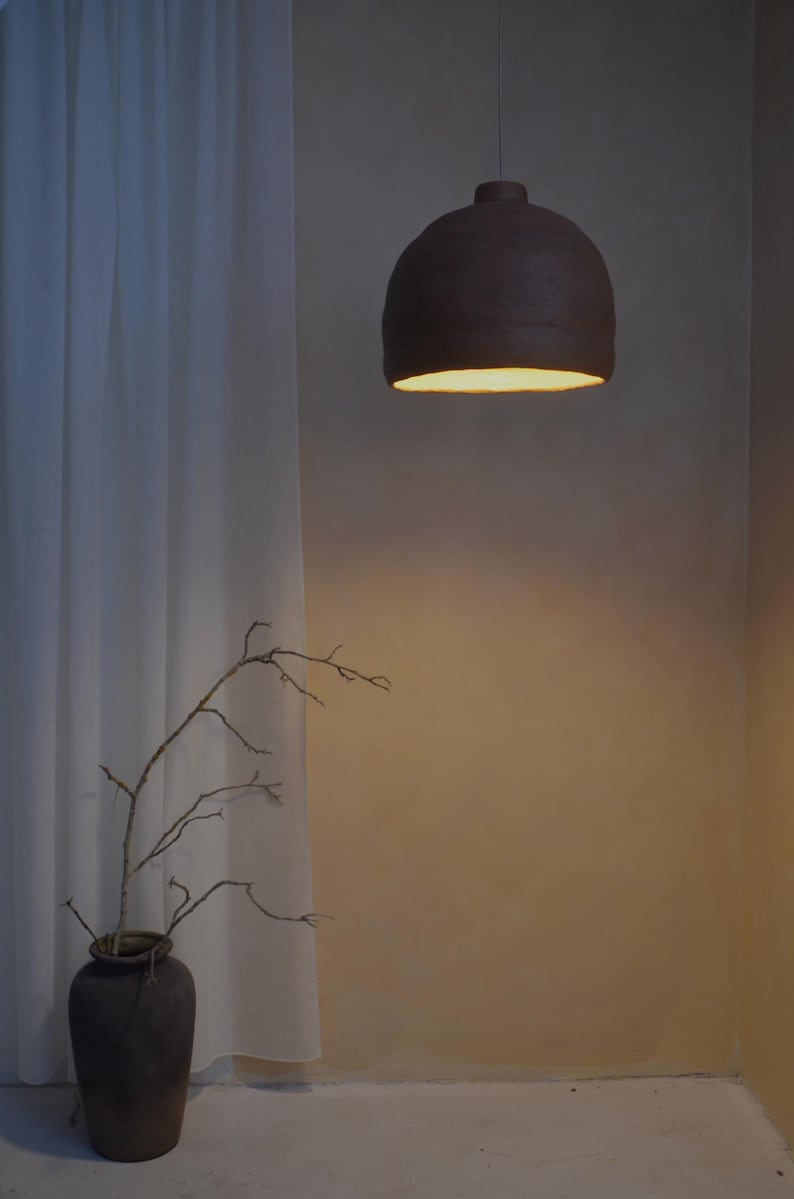 May include: A dark brown, dome-shaped pendant light fixture hangs from the ceiling. The light fixture is illuminated from within, casting a warm glow. A vase with branches is in the foreground, and a white curtain is to the left.