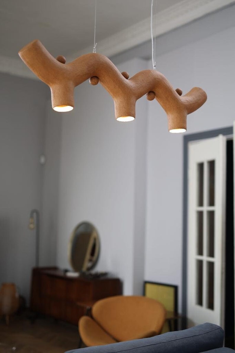 May include: A horizontal, branch-shaped pendant light fixture with a textured, light brown finish. The structure features three downward-facing cylindrical light sockets, each emitting a warm, bright glow, suspended by thin wires from the ceiling.