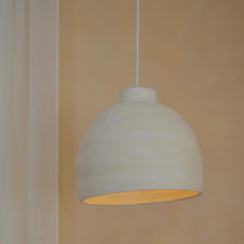 May include: Cream-colored, dome-shaped pendant light fixture. The light hangs from a white cord and has a textured surface. The interior of the lamp is illuminated with a warm, soft glow, creating a cozy ambiance.