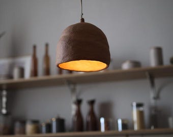 May include: A dome-shaped pendant light fitting with a textured, dark brown exterior and a warm, glowing interior. The lamp hangs from a thin cord, featuring a rough, organic surface finish that contrasts with the soft light emitted from below.
