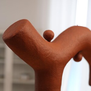 May include: A terracotta-colored sculptural object with a textured, matte surface. The form features a thick, branching structure with a cylindrical base and a smaller, spherical protrusion attached to the top of the main arm.