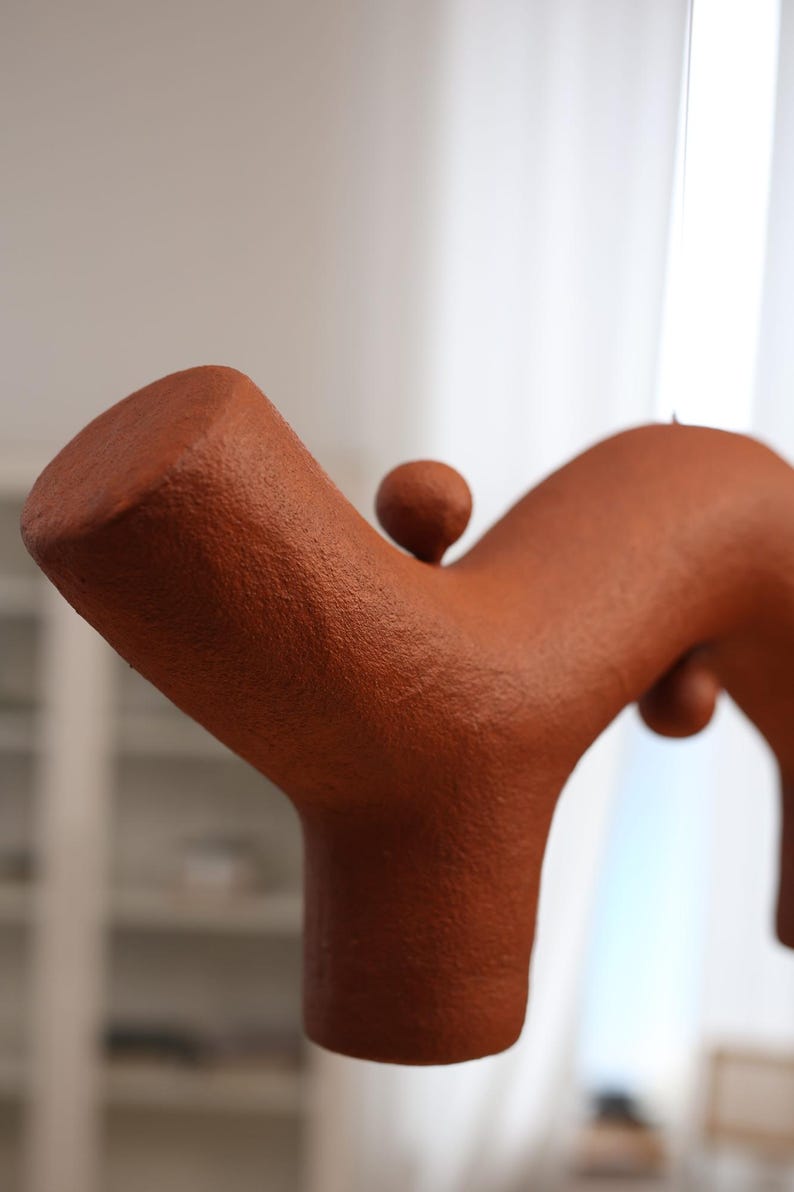 May include: A terracotta-colored sculptural object with a textured, matte surface. The form features a thick, branching structure with a cylindrical base and a smaller, spherical protrusion attached to the top of the main arm.