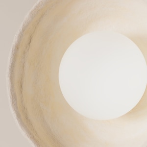 May include: A top-down view of a circular pendant light fitting featuring a wide, textured, off-white shade with an irregular, organic rim. A smooth, spherical white light bulb is positioned centrally within the shade, emitting a soft, even glow.