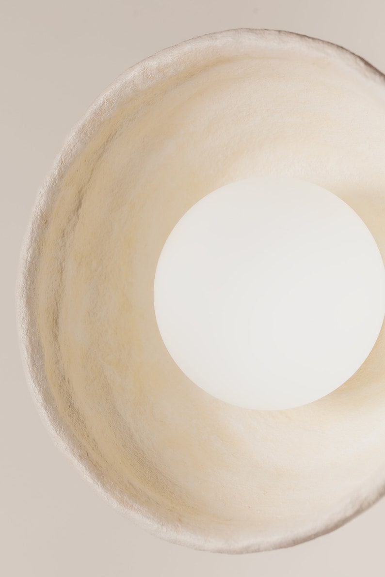 May include: A top-down view of a circular pendant light fitting featuring a wide, textured, off-white shade with an irregular, organic rim. A smooth, spherical white light bulb is positioned centrally within the shade, emitting a soft, even glow.