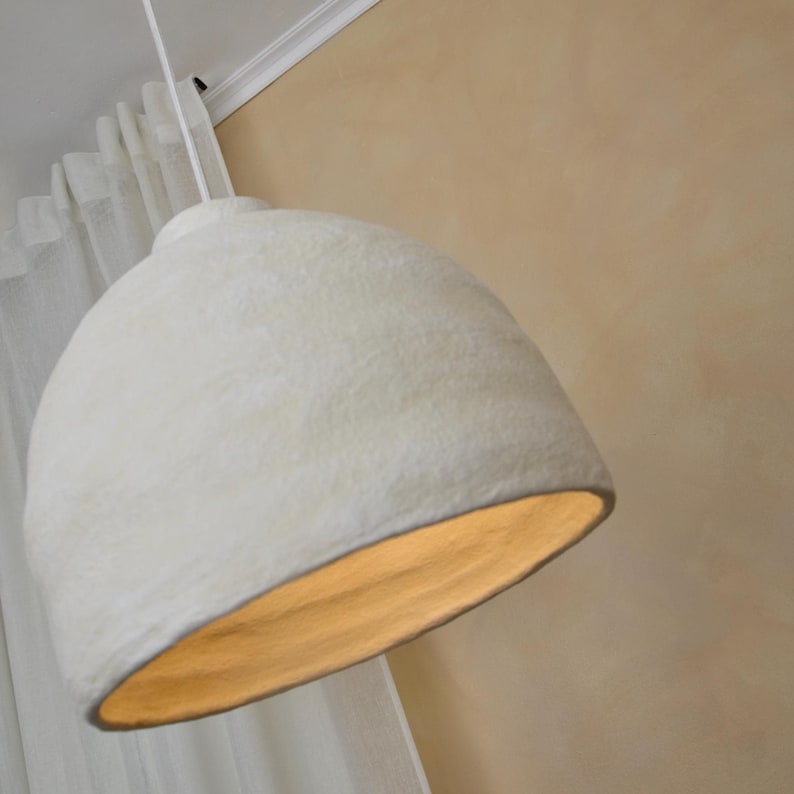 May include: A white, dome-shaped pendant light fixture. The light has a textured surface and a warm, golden glow emanating from the interior. The fixture is suspended from a white cord against a neutral-toned wall.
