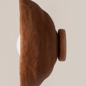 May include: A wall-mounted light fitting featuring a bowl-shaped shade with a rough, textured, dark brown terracotta finish. The fitting includes a circular base plate and a white spherical light bulb visible within the centre of the bowl.