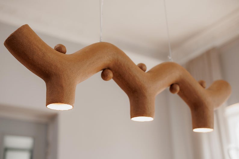 May include: A brown, textured, branch-shaped pendant light fixture with a wavy, organic form. The fixture features three downward-facing circular light openings and several small, spherical decorative protrusions along its length.