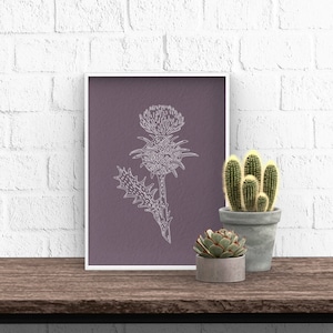 Puede incluir: Un dibujo de línea blanca de una flor de cardo sobre un fondo morado en un marco blanco. El marco está sentado en una mesa de madera con dos pequeñas suculentas en macetas.