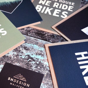 May include: A collection of greeting cards with various designs and text. The cards are on a rustic wooden surface. The text on the cards includes "All of the Plants", "We Ride Bikes", "Hike & Bike", and "SHDESIGN MALVERN".