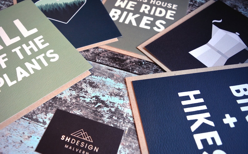May include: A collection of greeting cards with various designs and text. The cards are on a rustic wooden surface. The text on the cards includes "All of the Plants", "We Ride Bikes", "Hike & Bike", and "SHDESIGN MALVERN".