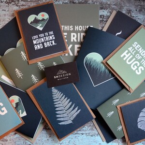 May include: A collection of greeting cards with various designs, including a mountain range, a fern, and a monstera leaf. The cards are all in shades of green and black with white text. The text on the cards includes "Love you to the mountains and back", "Sending all of the hugs", and "Paper hug".