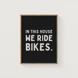 May include: Black framed sign with white text that reads "In this house we ride bikes."
