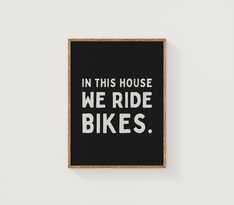 May include: Black framed sign with white text that reads "In this house we ride bikes."