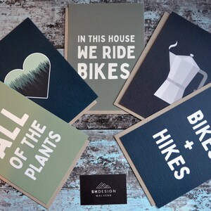 May include: Five greeting cards with different designs. The cards are all on a wooden surface. The cards have a variety of designs, including a heart with trees, a coffee pot, a bike, and a text design that says "In this house we ride bikes".