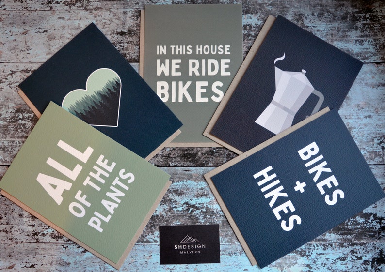 May include: Five greeting cards with different designs. The cards are all on a wooden surface. The cards have a variety of designs, including a heart with trees, a coffee pot, a bike, and a text design that says "In this house we ride bikes".