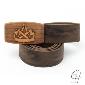 Könnte beinhalten: Ein brauner Ledergürtel mit einer Holzschnalle, die ein lasergraviertes Design aus zwei gekreuzten Äxten und einer Säge zeigt. Die Schnalle ist rechteckig und hat eine glatte, polierte Oberfläche.