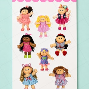 May include: A sticker sheet titled Daughter Lily Design featuring eight diverse dolls with yarn hair, round faces, and various outfits including dresses, a bear-themed onesie, and nautical attire, all displayed on a white background.