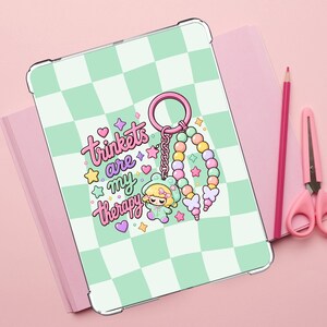 May include: A tablet case featuring a light green and white checkered pattern. The center displays a graphic of a keychain with colorful beads, a small character in a frog hood, and the text "trinkets are my therapy" surrounded by stars and hearts.