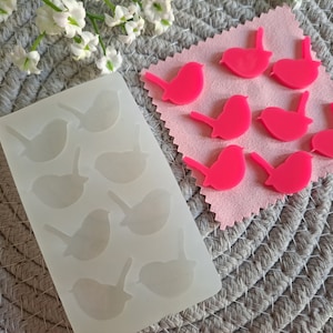 May include: A white silicone mould with 10 cavities shaped like small birds. The mould is on a grey textured surface. Next to the mould are 8 pink plastic birds.