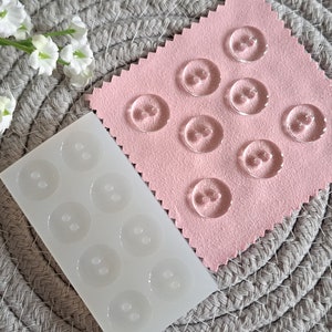 May include: A white silicone mould with nine circular cavities for making resin buttons. The mould is next to a pink fabric surface with nine clear resin buttons.
