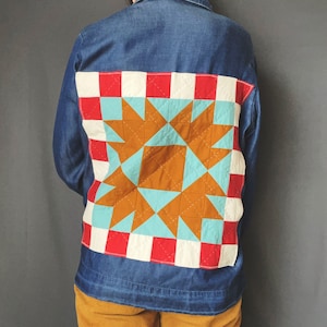 May include: A denim jacket with a patchwork quilt design on the back. The quilt is made of red, white, blue, and brown fabric and features a star pattern.