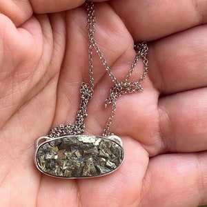 Könnte beinhalten: Eine silberfarbene Halskette mit einem rechteckigen Anhänger, der mit einem strukturierten Pyritstein gefüllt ist. Der Pyrit hat Gold- und Silbertöne. Die Kette ist filigran und der Anhänger ist in eine silberne Lünette gefasst. Der Text "DRAGONFLIESUNDBUTTERFLIES" befindet sich unten.