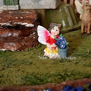 May include: A miniature fairy figurine with pink wings, a red dress, and a yellow skirt, holding a small pot of blue flowers. The fairy is standing on a bed of green moss.